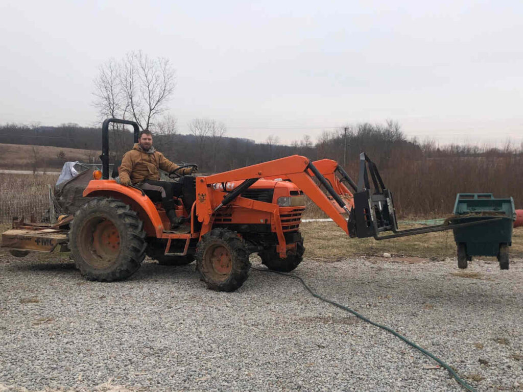 Tractor carrying small green wheelbarrow full of manure